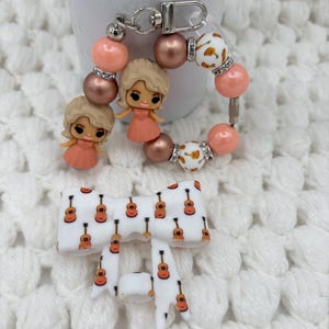 Puede incluir: Un llavero con dos dijes de muñecas rubias con vestidos color melocotón, intercalados con cuentas color melocotón, bronce metálico y blancas con estampado de guitarras y detalles plateados. Debajo, un lazo blanco con estampado de guitarras.