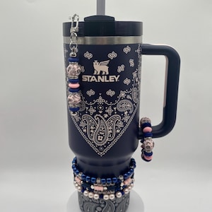 May include: Navy blue Stanley tumbler with a white paisley pattern and a black handle. The tumbler has a straw and is decorated with beaded bracelets in pink and blue. A beaded charm hangs from the lid.