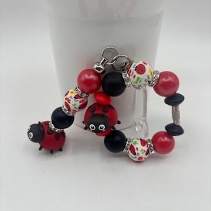 Op de afbeelding: Een armband met een lieveheersbeestjesthema met rode, zwarte en witte kralen. De armband heeft lieveheersbeestjesbedels en kralen met bloemen- en strassaccenten. De armband staat op een transparante standaard tegen een witte achtergrond.