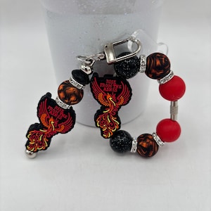 Peut inclure: Un porte-clés perlé avec deux breloques phénix en silicone noir portant l'inscription "RISE FROM THE ASHES" en rouge et orange. Le design comprend des perles rondes à paillettes noires, rouges et à motifs tourbillonnants orange et noirs.