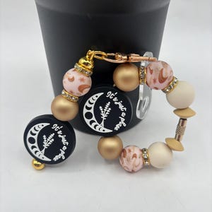 May include: A beaded keychain and matching round charm with the text "It's just a phase". The keychain features gold, pink, and cream-colored beads, with a gold clasp. The charm is black with a white crescent moon and floral design.