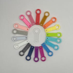 Può includere: Un set di 18 portachiavi colorati in varie tonalità di rosa, arancio, giallo, verde, blu, viola e grigio. I portachiavi sono in plastica e hanno un design semplice e arrotondato.