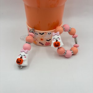 May include: A Halloween-themed beaded accessory featuring white ghost charms holding orange pumpkins, pink bows, and pink and orange beads. The accessory is shown next to a peach-colored tumbler.