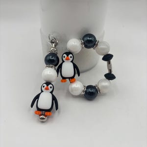 May include: A penguin-themed jewelry set featuring a bracelet and a charm. The bracelet has white and dark gray beads with rhinestone accents. The charm and bracelet both have small penguin figures with orange feet.