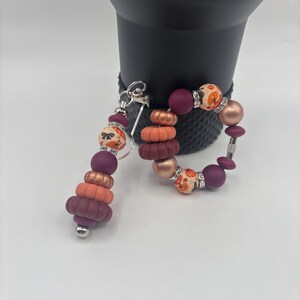 May include: A beaded keychain and bracelet set. The keychain features stacked beads in shades of burgundy, orange, and copper, with a silver-tone clasp. The bracelet has similar beads, including some with a floral pattern and rhinestone accents.