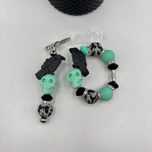 Op de afbeelding: Een set handgemaakte accessoires met een thema van raven en schedels. De set bevat een sleutelhanger en een armband. De sleutelhanger en armband hebben zwarte ravenbedels, mintgroene schedel kralen en zwart-wit patronen kralen.