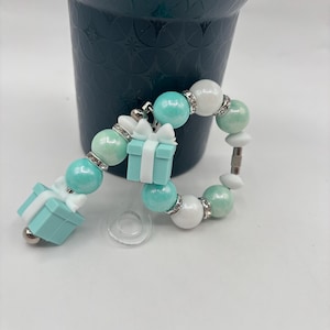 Może przedstawiać: Bransoletka z koralików z turkusowymi, białymi i jasnozielonymi koralikami, srebrnymi przekładkami i małymi zawieszkami w kształcie pudełka na prezent. Bransoletka została zaprojektowana z połączenia okrągłych i dekoracyjnych elementów.