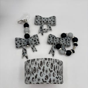 May include: A collection of Halloween-themed accessories. The set includes a bow, a bow with a clip, and a bracelet, all featuring a gray and black spooky pattern. A matching decorative band with Halloween icons is also included.