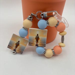 Peut inclure: Un bracelet de poignet à perles avec des breloques carrées représentant une image religieuse, entrecoupées de perles aux couleurs pastel. Le bracelet comprend des perles bleu clair, pêche et jaune, avec des accents argentés.