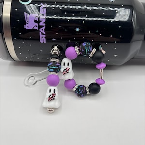 May include: A black Stanley tumbler featuring a celestial pattern with stars, a crescent moon, and a bat, paired with a beaded accessory. The accessory includes white ghost-shaped beads holding pink bats, purple and black matte beads, and silver spacers.