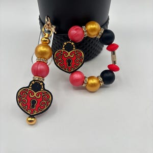 May include: A keychain and bracelet set featuring heart-shaped lock charms with red and gold filigree designs. The keychain and bracelet include gold and red beads, with rhinestone accents. The bracelet also has black beads.