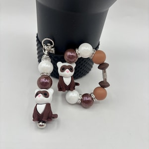 May include: A beaded keychain and bracelet set featuring ferret charms. The keychain has white, brown, and sparkly beads. The bracelet has white, brown, and sparkly beads, with a ferret charm. The ferret charms are brown and white.
