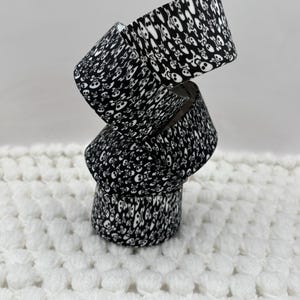 May include: A stack of three rolls of black and white adhesive tape featuring a repeating pattern of skulls and crossbones. The rolls are balanced vertically on top of each other, displaying the continuous graphic print across the surface of each roll.