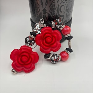 May include: Red rose-shaped earrings with black and white skull beads and silver accents. The earrings are attached to a silver clasp. The background features a black decorative cup with a gothic design.