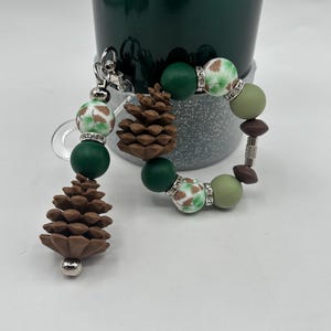 May include: A beaded keychain and bracelet set. The keychain has a brown pinecone charm and green and white patterned beads. The bracelet has similar beads in shades of green and brown. Both have silver-tone accents.