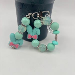 Puede incluir: Un llavero de pulsera con cuentas verde menta con un dije de conejito. La pulsera presenta cuentas redondas, cuentas de acento brillantes y un cierre plateado. El dije de conejito tiene un lazo rosa. La pulsera está junto a una maceta decorativa verde oscuro.