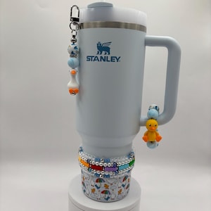 Puede incluir: Un vaso Stanley azul claro con tapa y asa blancas. El vaso está decorado con un charm de cuentas y un charm de pato. La base del vaso tiene un estampado de patos y paraguas.