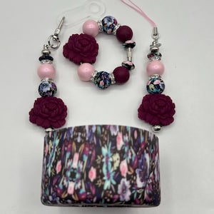 Op de afbeelding: Een set accessoires met een bloemig thema. Inclusief een armband, twee kralenbedels en een bekerwikkel. De armband en bedels hebben roze en bordeauxrode kralen, bloemaccenten en bordeauxrode bloemvormige bedels. De bekerwikkel heeft een donker bloemenpatroon.