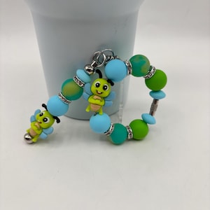 Op de afbeelding: Een kralenarmband en sleutelhanger met een bijen thema. De armband en sleutelhanger hebben blauwe, groene en gele kralen, met zilveren accenten. Elk stuk heeft een kleine, lachende bijenbedel.