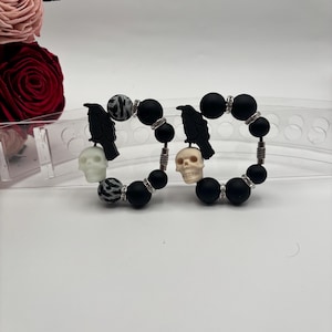 Op de afbeelding: Twee zwarte kralenarmbanden met een schedel- en ravenhanger. De kralen zijn zwart met zilveren accenten. De hangers zijn wit en zwart.
