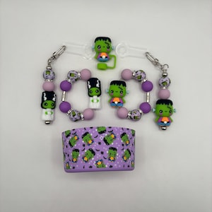 May include: A collection of Halloween-themed accessories featuring a Frankenstein's monster design. Includes beaded keychains, bracelets, and a purple band with a repeating monster pattern. The accessories have green, purple, and orange accents.