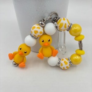 Peut inclure: Un porte-clés circulaire composé de perles jaunes et blanches, dont certaines présentent un motif de petits canards jaunes. Deux breloques canards jaunes en trois dimensions avec becs et pattes orange sont fixées à l'anneau avec des entretoises argentées.