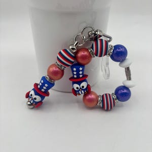 May include: A patriotic keychain featuring red, white, and blue beads, including striped and solid spheres. Two owl charms wear Uncle Sam hats. The keychain is attached to a silver-colored clasp.