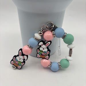 May include: A beaded bracelet with an Easter theme, featuring a cartoon bunny charm carrying a basket of eggs. The bracelet includes pink, blue, green, and white beads, with silver accents. The bunny charm is black with colourful details.
