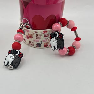 May include: A pink and red beaded keychain with a charm featuring two hugging cats. The keychain is attached to a pink and white mug with a cat pattern. The mug is next to a larger pink cup.