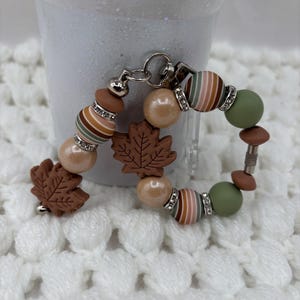May include: A beaded keychain featuring two brown maple leaf charms, round beads in shades of olive green, tan, and striped earth tones, and silver-toned metal spacers with clear crystals. The beads are arranged on a circular metal ring.
