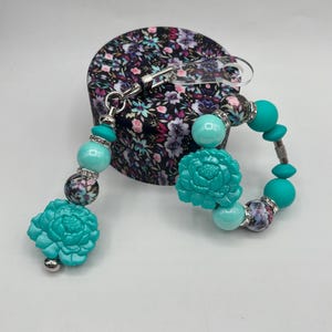 Może przedstawiać: Turkusowa bransoletka z koralików i pasujący brelok do kluczy z kwiatowymi akcentami. Bransoletka i brelok do kluczy mają duże turkusowe koraliki w kształcie kwiatów, okrągłe turkusowe koraliki i koraliki we wzory kwiatowe. Przedmioty są wyświetlane w pobliżu pudełka z kwiatowym wzorem.