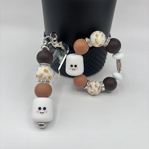 May include: A beaded keychain and bracelet set. The keychain features a series of brown, white, and speckled beads, with a smiling marshmallow charm. The bracelet has similar beads and a matching marshmallow charm. Both have silver-tone hardware.