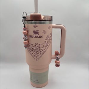 Peut inclure: Un gobelet Stanley Quencher H2.0 FlowState rose clair avec une poignée, une paille et des breloques perlées décoratives. Le gobelet présente un motif bandana floral et le logo Stanley. La poignée a un charme perlé assorti.