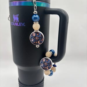 Puede incluir: Vaso Stanley negro con un charm plateado. El charm presenta un diseño celestial con lunas y estrellas sobre un fondo azul oscuro, acentuado con cuentas azules y beige. El vaso tiene un asa negra y una base texturizada.