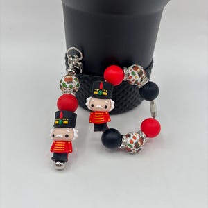 May include: A Christmas-themed keychain and bracelet set. The keychain features a silver clasp and a charm of a nutcracker soldier. The bracelet has red, black, and patterned beads, and a nutcracker charm.