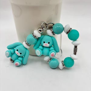 Könnte beinhalten: Zwei türkisfarbene Hasenanhänger mit rosa Akzenten und ein Armband mit türkisfarbenen und weißen Perlen. Die Anhänger und das Armband haben silberfarbene Metallakzente. Die Hasenanhänger haben ein niedliches, cartoonartiges Design.