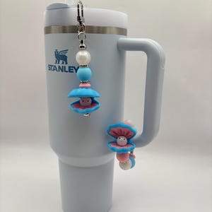 May include: A light blue Stanley tumbler with a white rim and handle, featuring a seashell-themed charm. The charm includes a blue shell, a pink pearl, and a silver clasp. A second charm is attached to the tumbler handle.