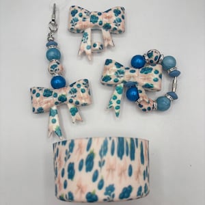 May include: A collection of handmade accessories featuring a floral bow design in shades of blue and pink. The set includes a keychain, earrings, and a bracelet with blue beads, and a matching cylindrical container.
