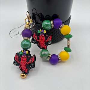 Puede incluir: Un juego de llavero y pulsera con temática de Mardi Gras. El llavero presenta cuentas moradas, verdes y doradas con un dije de cangrejo rojo. La pulsera tiene cuentas similares y un dije de cangrejo a juego.