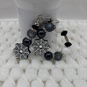 May include: A circular beaded keychain featuring black and white spiderweb-patterned beads, solid black and white spherical beads, and two black spiderweb-shaped charms with white spider graphics, all connected by silver-toned metal hardware.