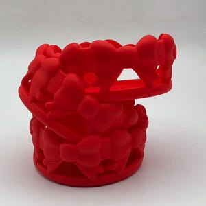 May include: A bright red silicone cup sleeve featuring a repeating pattern of small, raised bows. The cylindrical accessory has an open-work design with a continuous spiral structure, providing a textured grip for a standard-sized drinking tumbler.
