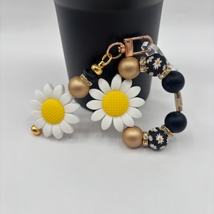 May include: A daisy-themed keychain wristlet with gold and black beads. The wristlet features white daisy charms with yellow centers, gold beads, and black beads with a white floral pattern. A gold clasp is attached.