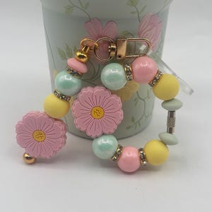 Può includere: Un portachiavi di perline dai colori pastello. Il portachiavi include ciondoli a forma di fiore rosa, perline gialle e rosa e perline blu iridescenti. Dettagli dorati e strass aggiungono brillantezza.