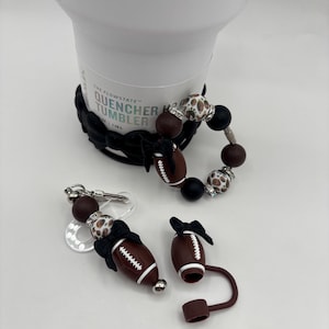 May include: A white tumbler with a black handle and a beaded bracelet. The bracelet has brown, black, and leopard print beads. There are also football-shaped accessories with black bows and a keychain with a football charm.