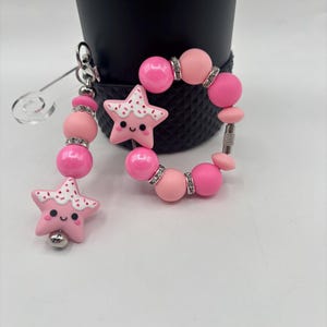 May include: A pink beaded keychain and bracelet set. The keychain features pink beads, a star charm with a smiling face, and a silver clasp. The bracelet has pink beads and rhinestone accents.