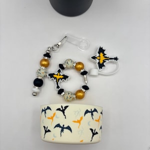 Può includere: Una collezione di accessori a tema pipistrello. Include un portachiavi con perline, un braccialetto, un tappo per bottiglia e una custodia per tazza. I disegni dei pipistrelli sono neri e gialli, con accenti dorati e bianchi. La custodia della tazza ha un motivo a pipistrelli ripetuto.