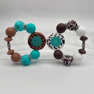 Könnte beinhalten: Zwei Armbänder mit Perlen in Türkis, Braun und Weiß. Die Armbänder haben runde Perlen, dekorative Mittelstücke und silberfarbene Akzente. Die Mittelstücke haben ein florales Design.