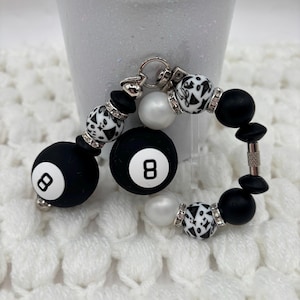 May include: A beaded keychain featuring two round black beads with white number eight designs, interspersed with smaller white beads, black matte beads, and patterned white beads with black geometric shapes, accented by silver-toned rhinestone spacers.