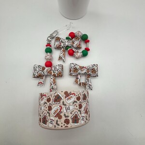 May include: A Christmas-themed set featuring a cup holder, bow-shaped hair clips, and a beaded keychain. The cup holder and bows have a repeating pattern of gingerbread men, houses, and milk bottles. The keychain and clips have red, green, and white beads.