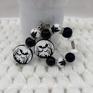 May include: A beaded keychain featuring round black glitter beads, white beads with black silhouettes of haunted houses, bats, and tombstones, and circular charms with a bat and graveyard scene. The beads are accented with small silver-toned rhinestone spacers.
